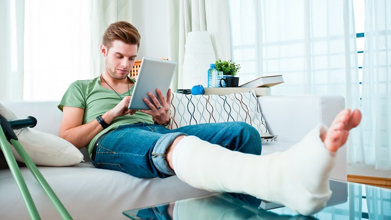 Grafika ilustracyjna przedstawiająca mężczyznę z nogą gipsie, który korzysta z tabletu.